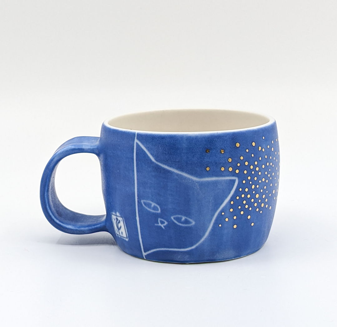 White mug with the outside painted blue. Decorated with gold dots and large simple drawing of a cat's head peeking around a wall.