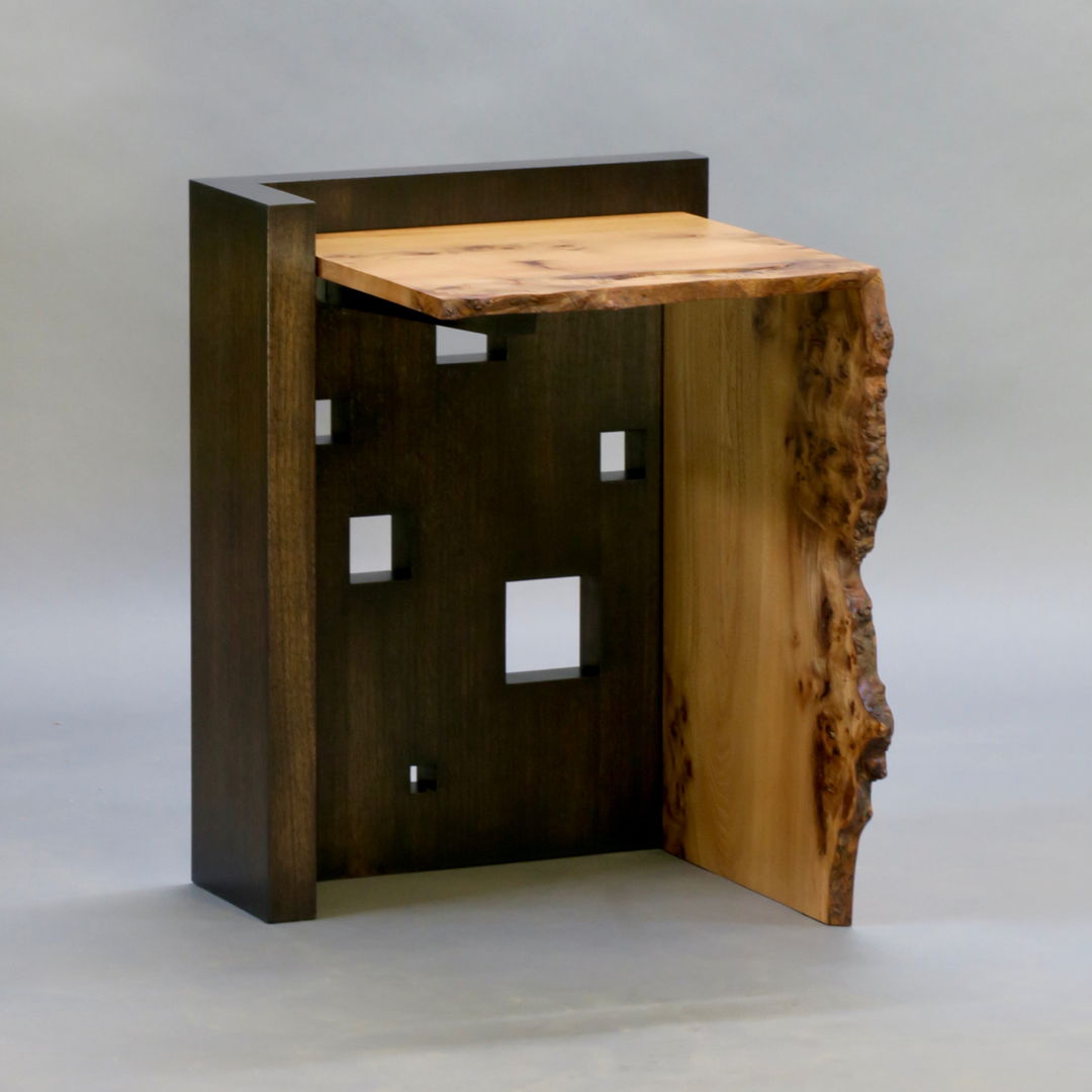 The Oriel IV side table features a blackened solid walnut base which has been perforated with square "windows" of various sizes creating a sculptural play of light and shadow. The live edge cantilevered top and waterfall joined leg in highly figured elm complete the structure.