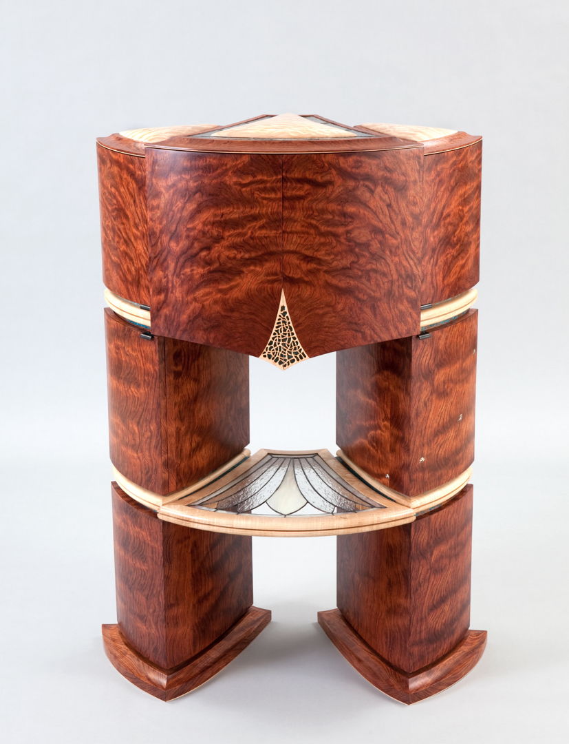 This complex fan-shaped piece  features three curved-sided irregular quadrilateral cabinets nested together and resting on a matching base. The exterior features waterfall bubinga  blistered big-leaf maple  copper-foiled art glass and mother of pearl inlays.The interior reveals patinated blue copper  mother of pearl and abalone inlays  a revolving carousel  pivoting drawers with pierced and carved bottoms and dividers  marquetry and art glass shelving.