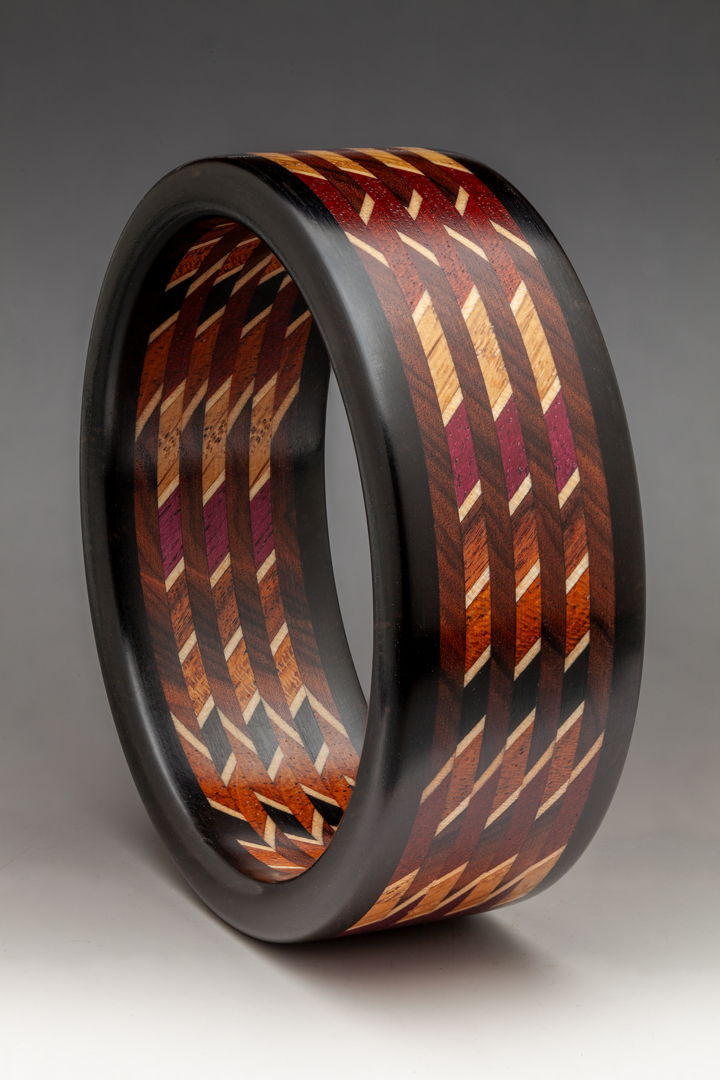 This dramatic and elegant linear Mosaic Bracelet is constructed with 12 different exotic and domestic sustainable hardwoods and Maple veneer. The laminate is cut on the bias a 45 degree angle cut and then the slices are stacked and orientated in a 90 degree rotation captured with Ebony edges.
