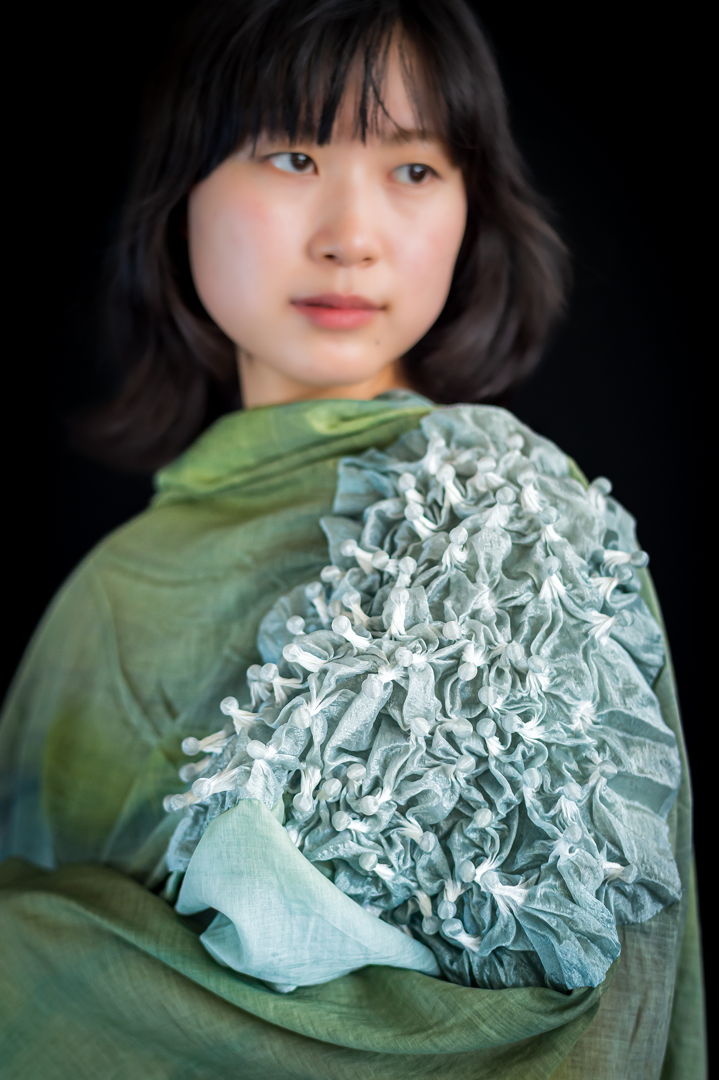The gray-ish green tones  reminiscent of shadows at the bottom of the sea  sparkle vividly in the flickering wind and glistening sunlight. The blend of silk and cotton fabric creates a beautiful layering of colors  producing complex sparkles with each drape. The combination of this fabric and the shibori dyeing technique adds a unique surprise and complexity to the beauty of the shawl.