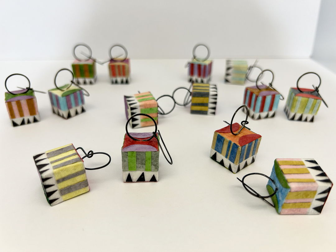 Circus Earrings are lightweight  cube-shaped earrings. Their dangle is made from annealed steel wire hammered and shaped into circles. The cubes are hollow and made from mat board  with layers of assorted colorful papers “painted” on with brush and glue. The same process is used for the colorful and detailed patterns. All paper jewelry is sealed for wear.
