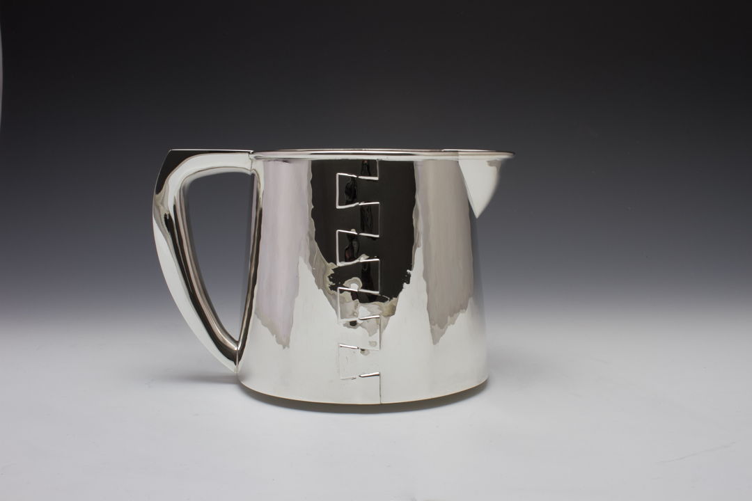 An elegant silver creamer with a constructed design that features a dovetail seam up either side of the piece and flowing hollow handle.