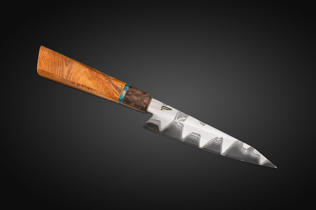 A kitchen Petty (utility knife) in San Mai construction. Blade is diffusion bonded with a 1095 high carbon core and 304 stainless on the sides. The handle is Greater Rhododendron with a Turkish Walnut bolster and a surf stone accent.