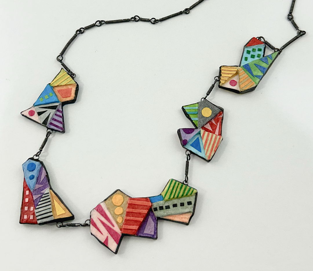 The Origami Rainbow Necklace is suggestive of the folds and shapes of origami. Each component is a geometric-shaped paper sculpture. Layers of mat board form multiple levels of geometric shapes for each component. Paper colors are “painted” on with brush and glue  as are the detailed designs. Hardware is wire-wrapped annealed steel links.