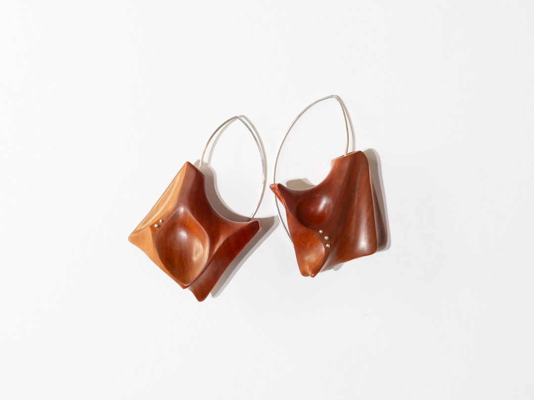 Hand shaped and polished Pink Ivory wood with Argentium Silver details and ear wires. A locking system is employed to bond the metal and wood. A keyhole system is drilled within the wood and the metal is knurled before epoxying wood and metal together.