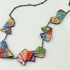 The Origami Rainbow Necklace is suggestive of the folds and shapes of origami. Each component is a geometric-shaped paper sculpture. Layers of mat board form multiple levels of geometric shapes for each component. Paper colors are “painted” on with brush and glue  as are the detailed designs. Hardware is wire-wrapped annealed steel links. 